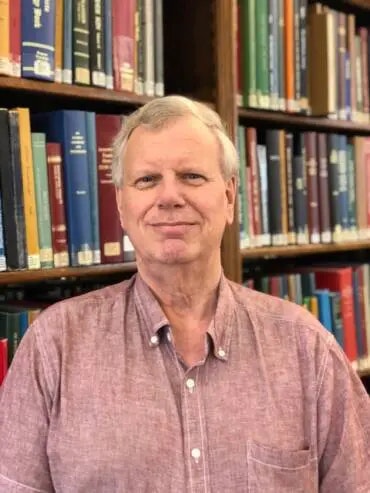 A man in a pink shirt smiling in front of bookshelves.