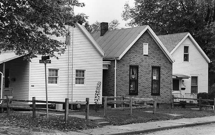 Two small houses with front porches, one with siding and the other brick, in a residential area. Black and white photo.