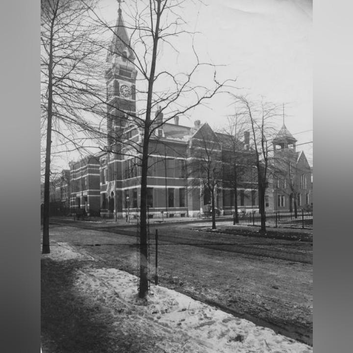 Historic building with clock tower, bare trees, snow patches, and park benches. Black and white photo.
