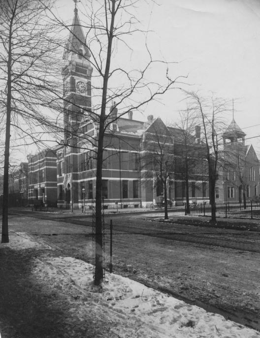 Historic building with clock tower, bare trees, snow patches, and park benches. Black and white photo.