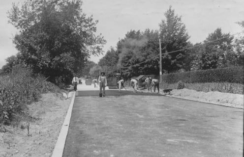 A vintage photo of people and horses on a road with trees and hedges.