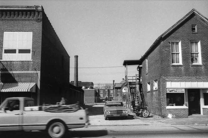Black and white photo of a street with a moving pickup truck, parked cars, brick buildings, and a fire escape.