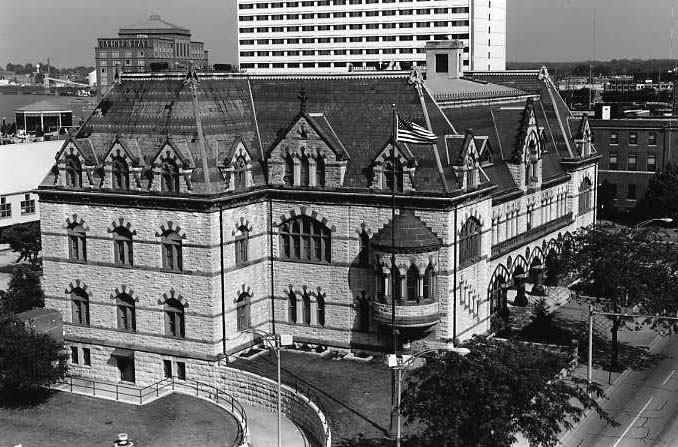 Historic stone building with architectural details, surrounded by modern buildings under a clear sky.