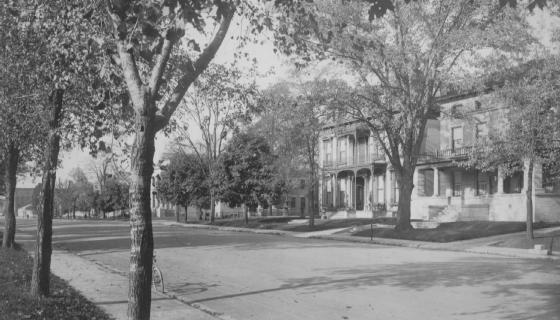 Vintage photo: tree-lined street with houses, no people visible, sepia toned, possibly early 20th century.