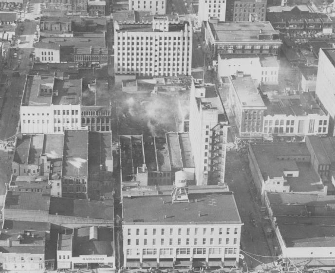 A black and white aerial view of urban buildings, possibly from mid-20th century, with visible smoke in the center.