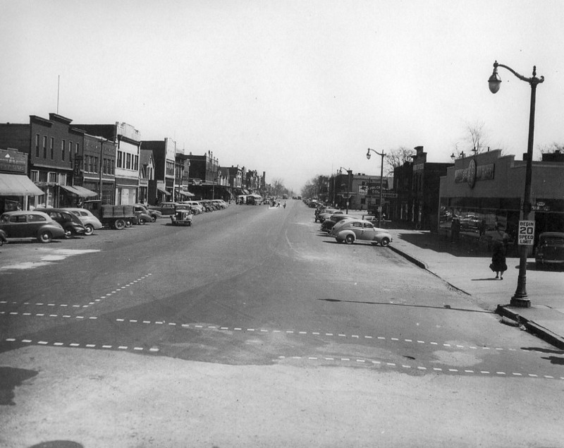 Vintage street scene with old cars, buildings, and a pedestrian.