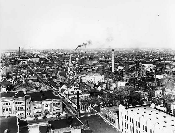 Historic black & white photo of a dense urban area with industrial smokestacks and a church spire.