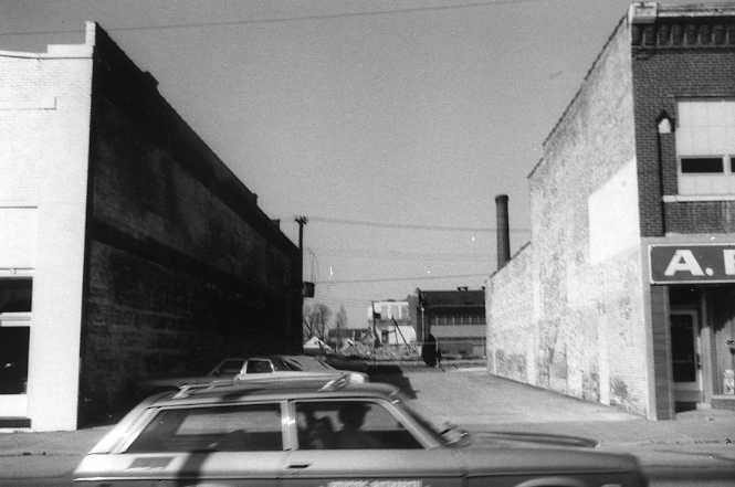 Black-and-white photo of an urban alley with cars and older buildings.