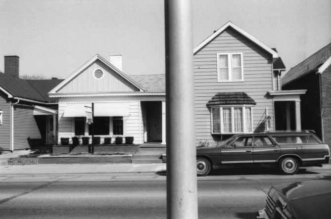 Black and white photo of suburban street with two houses and a vintage station wagon.