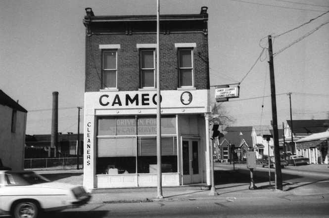 Black and white photo of an old building with a sign "CAMEO CLEANERS", a blurred car passing by, and a clear sky.