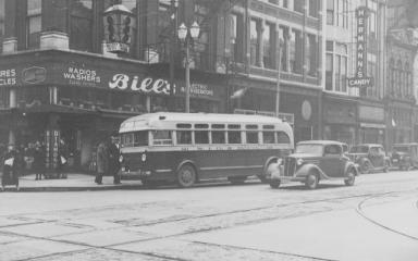 Vintage street scene with an old bus and cars, storefronts in the background. Black and white photo.