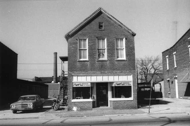 Black and white photo of a two-story brick building with a storefront, an old car parked outside, and a clear sky.
