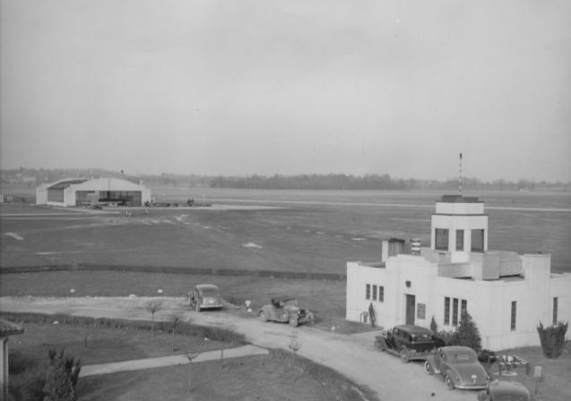 Vintage photo of an airfield with control tower, hangar, vintage cars, and open fields.