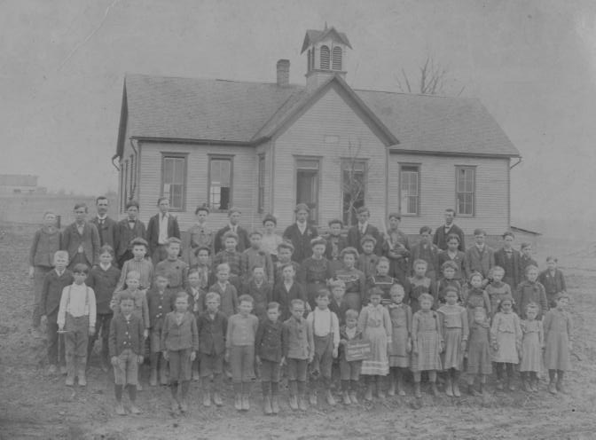 A group of people, likely a school class with teachers, standing in front of an old schoolhouse.
