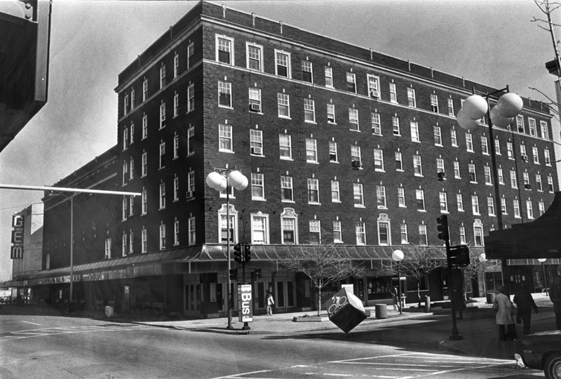 Black & white photo of a multistory building, street lamps, clear sky, pedestrians, and a parked car.
