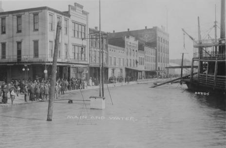 Historic flood scene with people, submerged streets, buildings, and a steamboat.