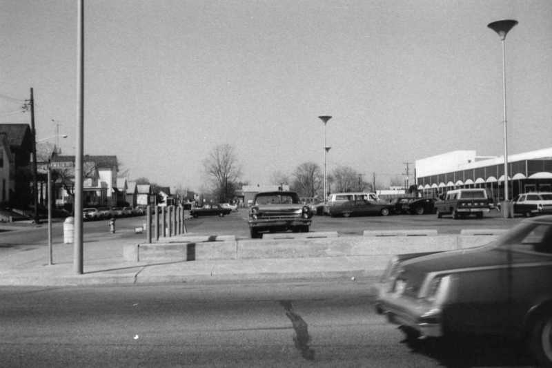 Black and white photo of a street with vintage cars, streetlights, and buildings, captured with a moving car in the foreground.