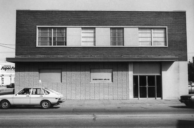 A two-story building with a brick facade, large windows on the upper floor, a parked car, and clear skies.