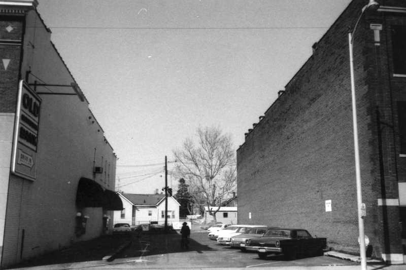 Black and white photo of a street with cars, buildings, and a person.