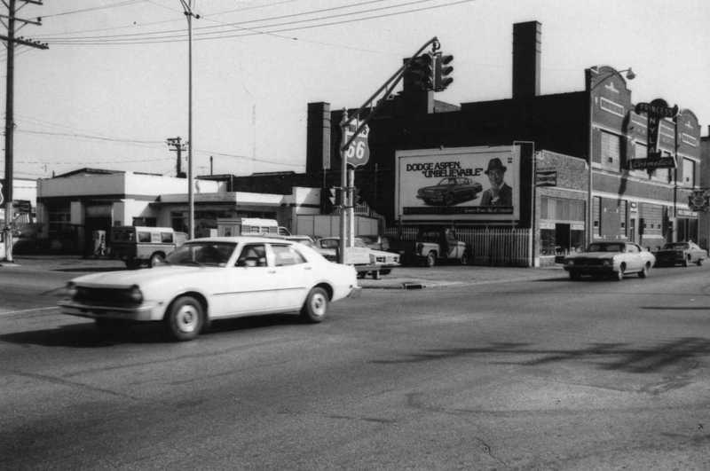 Street corner with cars, buildings, and a billboard for Dodge Aspen. Black and white photo.
