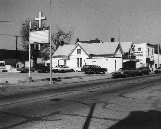 Black and white photo of a street scene with houses, cars, a church sign, and a business named "DORISS."