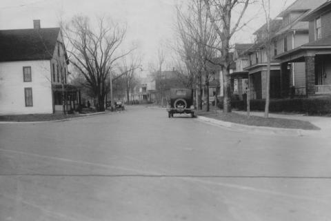 A vintage black-and-white photo of an old car on a residential street with houses and leafless trees.