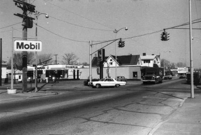 A black-and-white photo of a street corner with a Mobil gas station, traffic signals, cars, and other buildings.