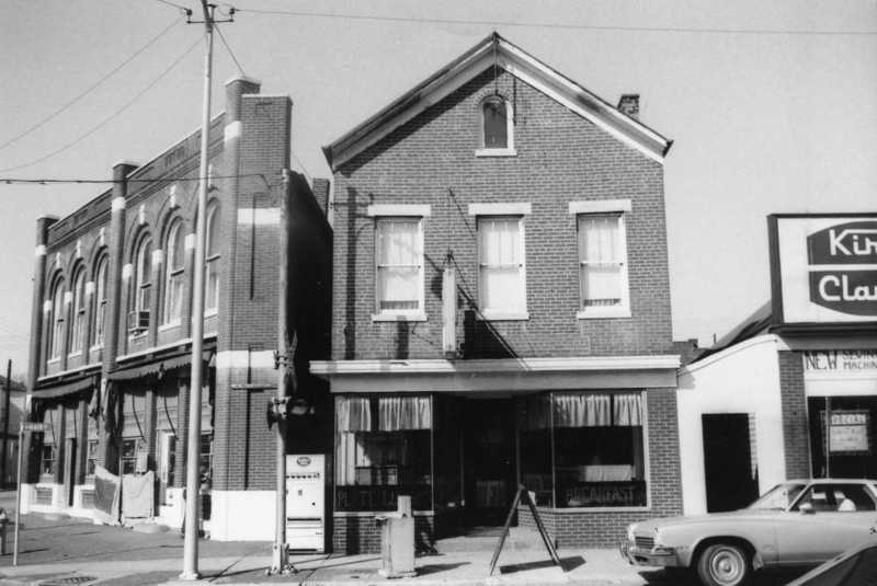 Vintage black and white photo of an old street with brick buildings, a classic car, and storefronts.