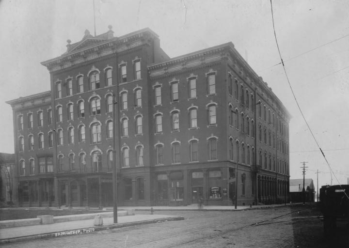 Historic building at a street corner with tram lines and an old car visible; black and white photo.
