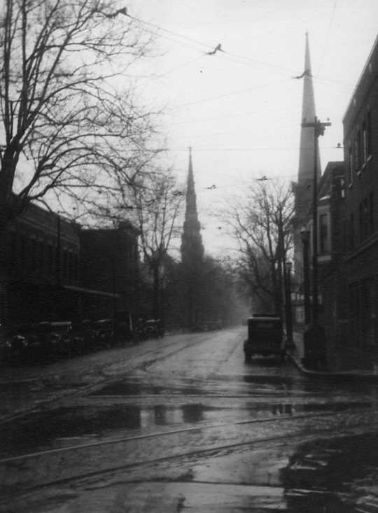Old street with tram tracks, vintage cars, church spire in distance, wet reflective surface; early to mid-20th century feel.