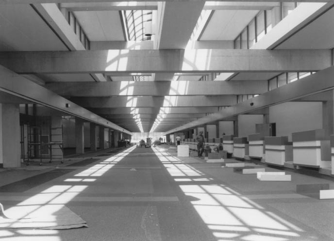 Interior of a large, empty building with exposed beams, skylights, and construction materials on the floor.