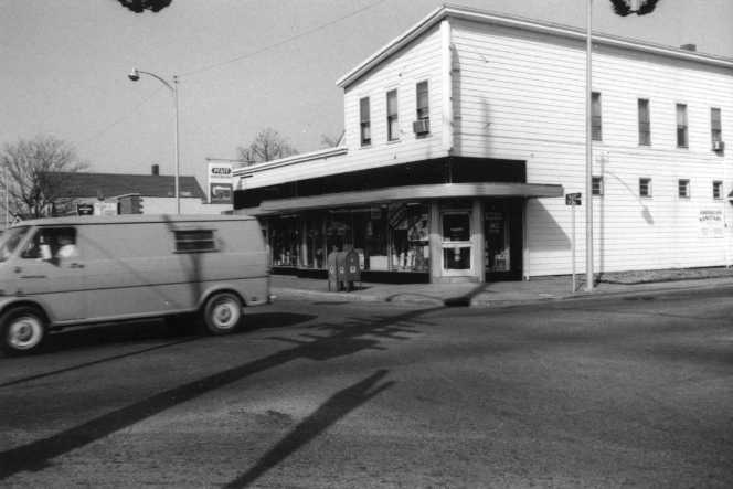 Black and white photo of a vintage van driving past a corner building with a covered walkway.