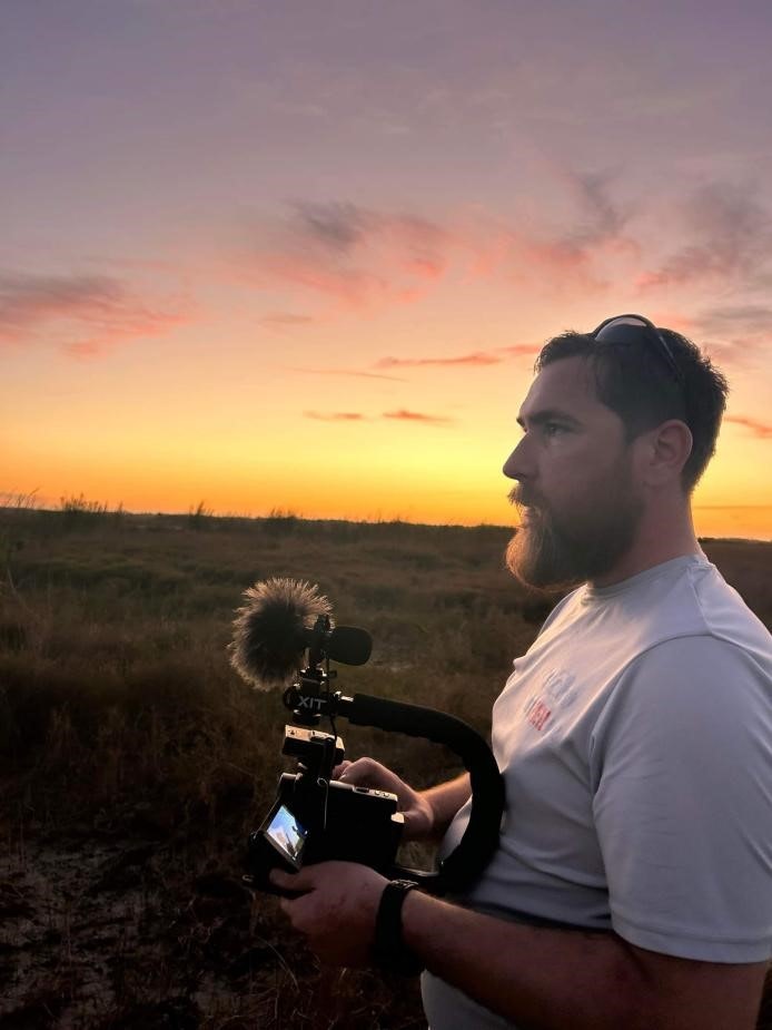 A person with a beard and sunglasses is holding a camera with a microphone, set against a sunset background.