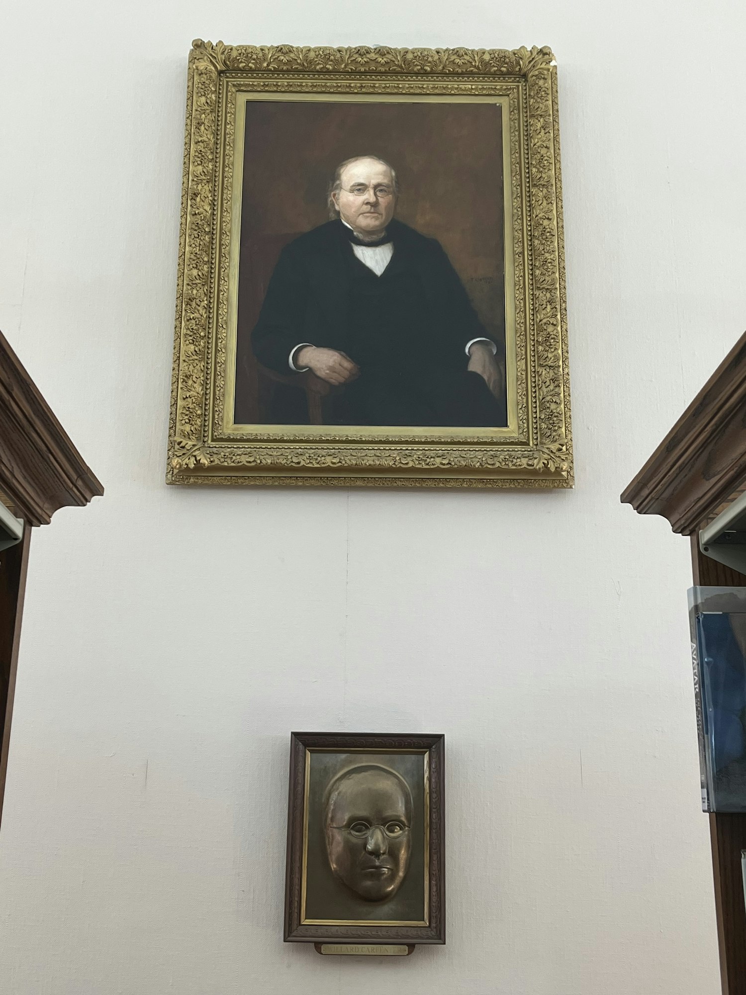 A framed portrait of a man in a suit, and a framed bronze mask on a white wall.