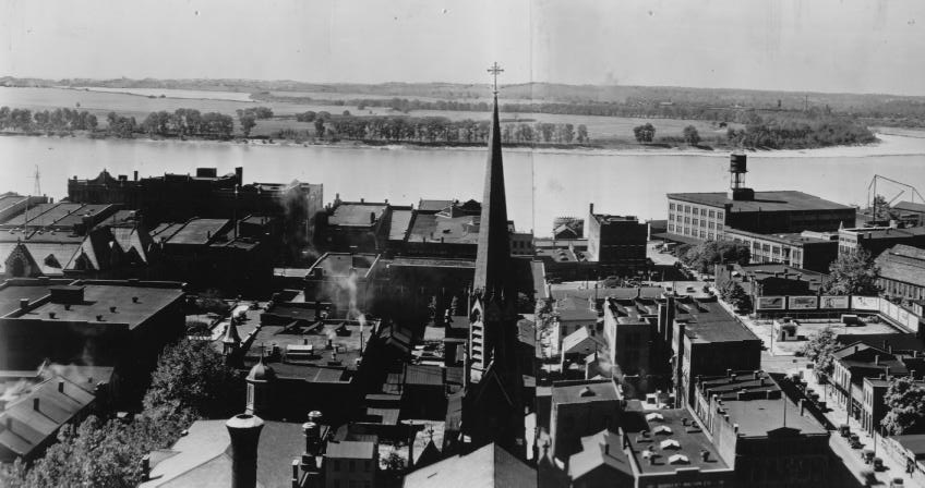 Historic town overlooking a river with a church spire, industrial buildings, and smokestacks. Black & white photo.