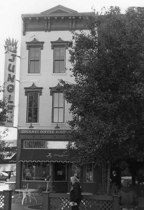 A black and white photo of a building with a sign "The Jungle Gourmet Coffee House," a tree, and a person walking by.