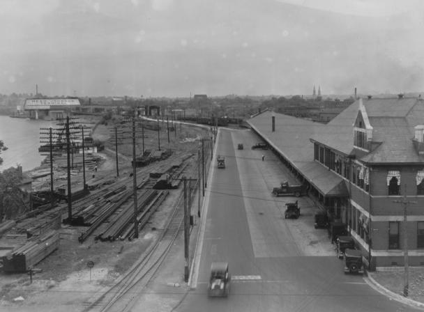 Vintage photo of a street with cars, railroad tracks, industrial buildings, and a river with a boat in the background.