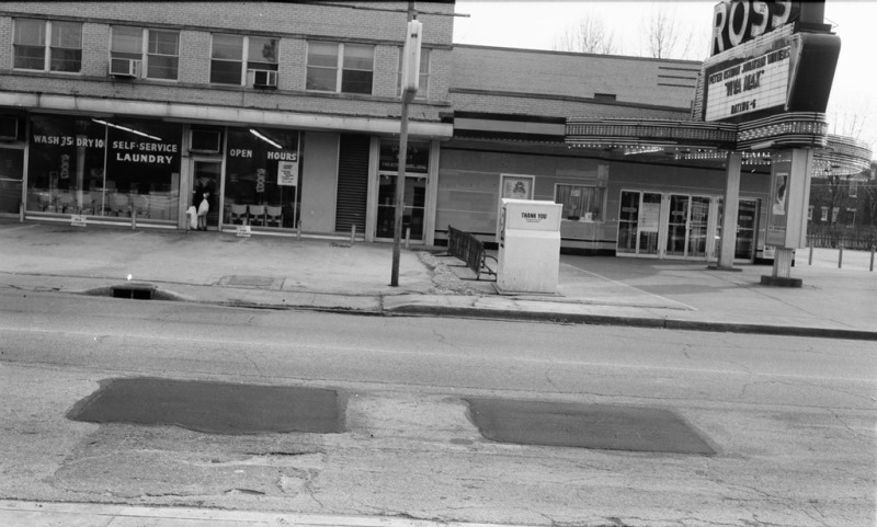 An old street view with a laundromat, a closed theater marquee, a bench, and patched asphalt.