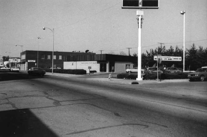 Black and white photo of a street corner with buildings, cars, and a tall signpost.