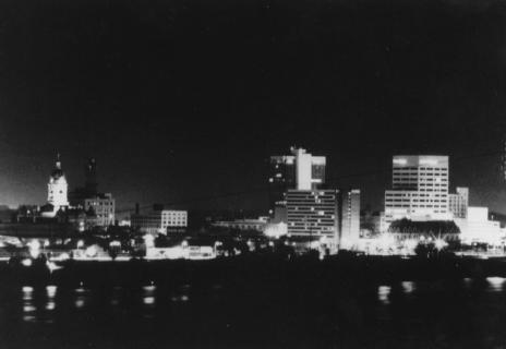 A black and white photo of a city skyline at night, with reflections on water.