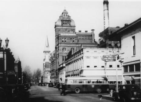 Historic street view with vintage cars, buildings with ornate architecture, and a tall smokestack. Black and white photo.