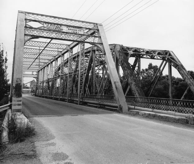 An old truss bridge with intricate metalwork, over a road, in black and white photography.