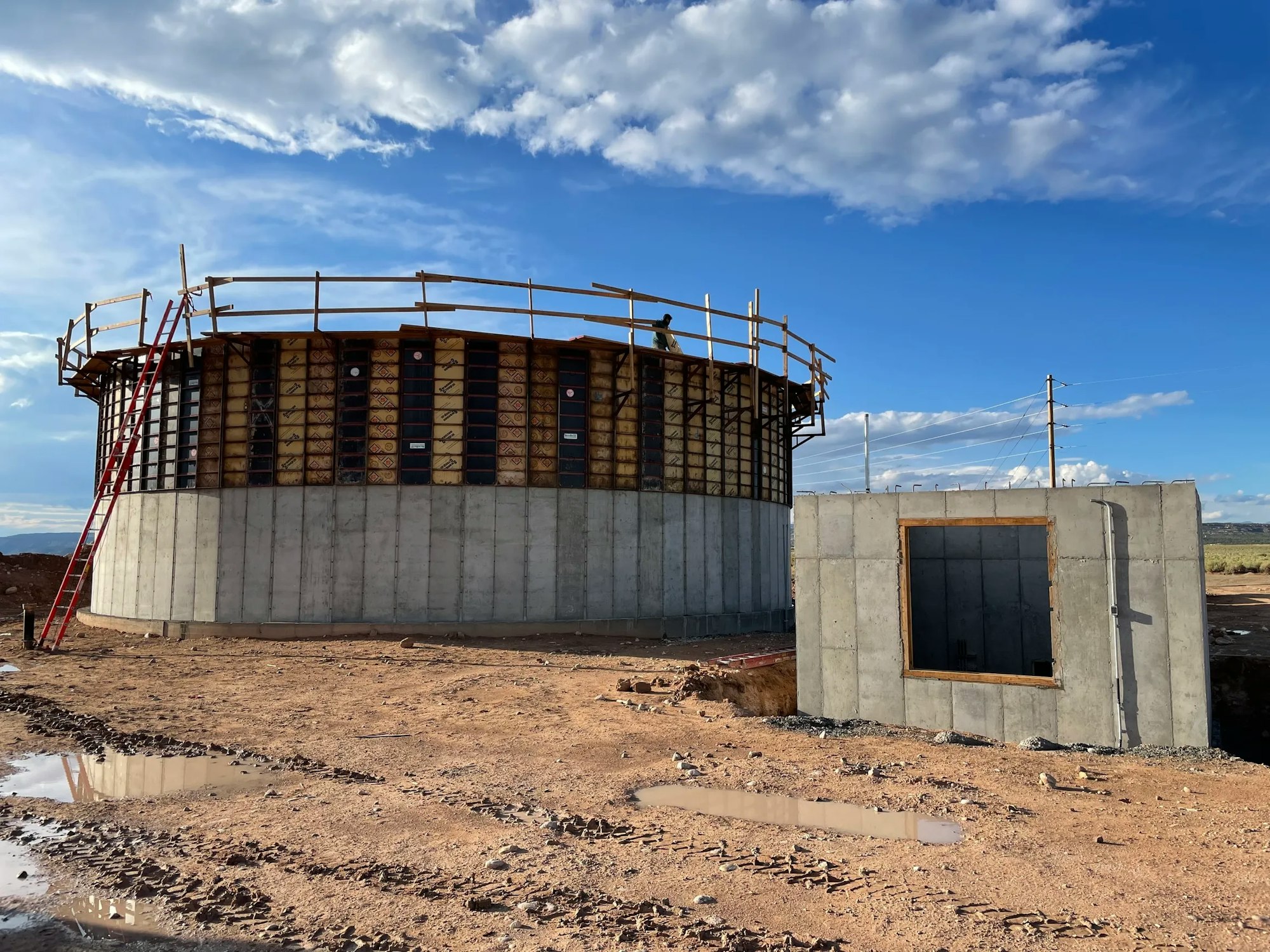 Construction site with a circular concrete structure and a wall with a rectangular opening under a blue sky.