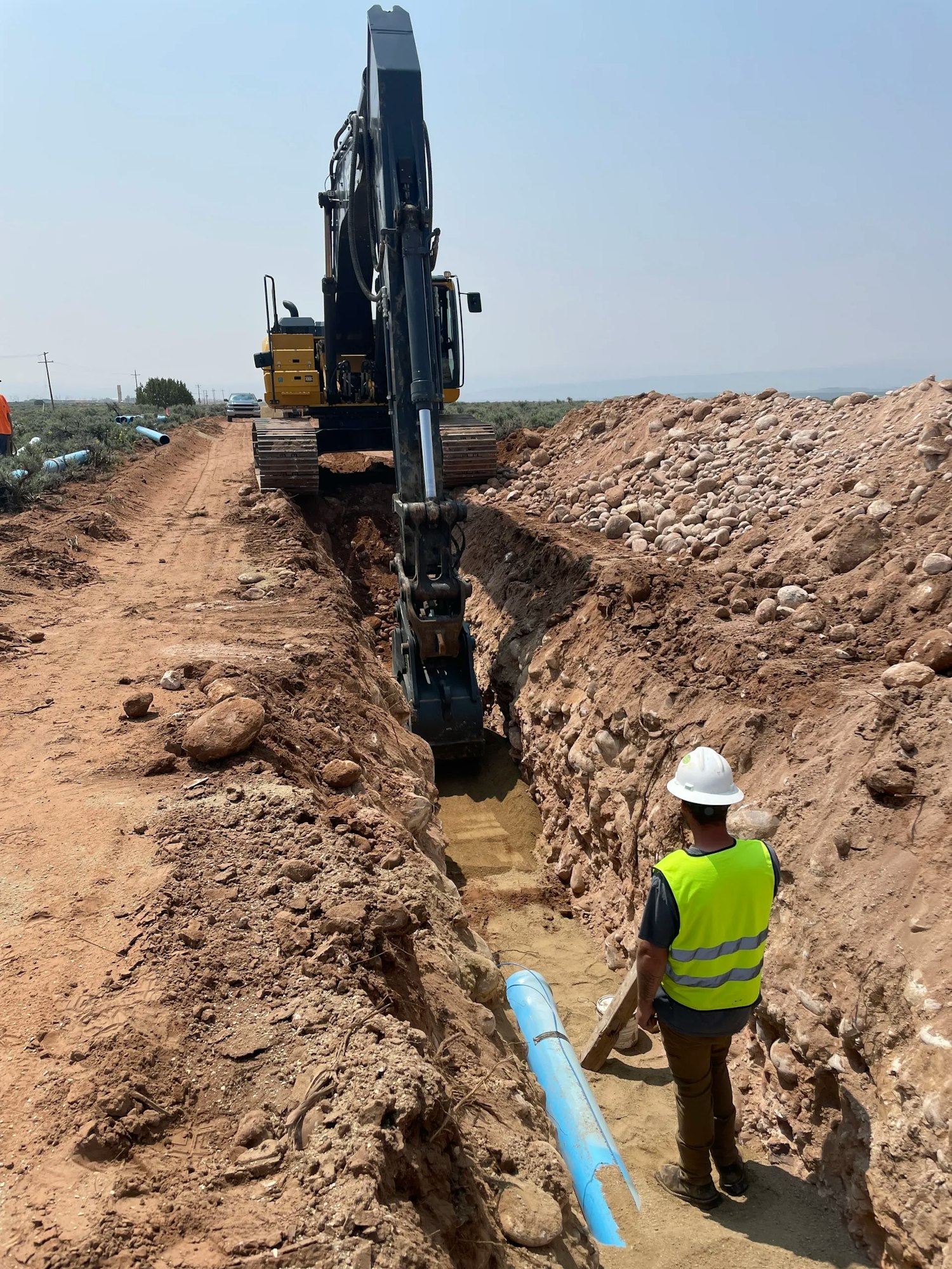 Excavator digging a trench with a worker in a safety vest laying blue piping.