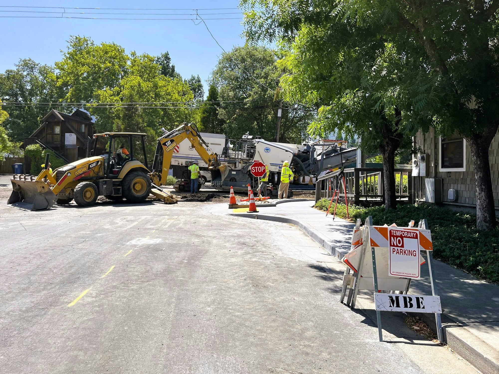 Pavement construction site with machinery, workers, and "Temporary No Parking" signs on a tree-lined street.