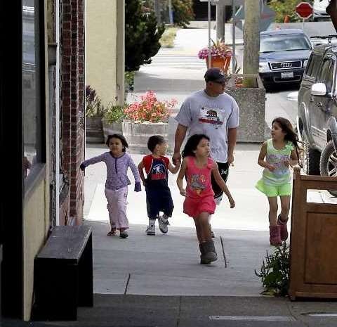 A man walks with four children on a sidewalk, smiling and enjoying a sunny day, with plants and parked cars around.