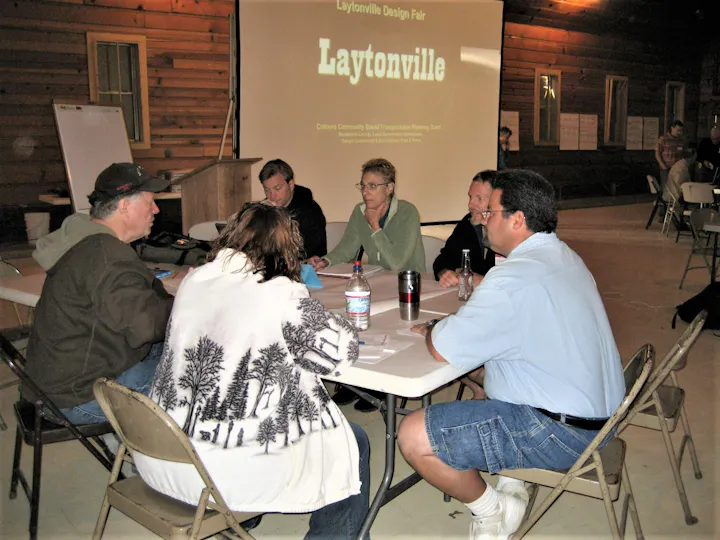 A group of people seated around a table during a community design fair in Laytonville, with a presentation in the background.