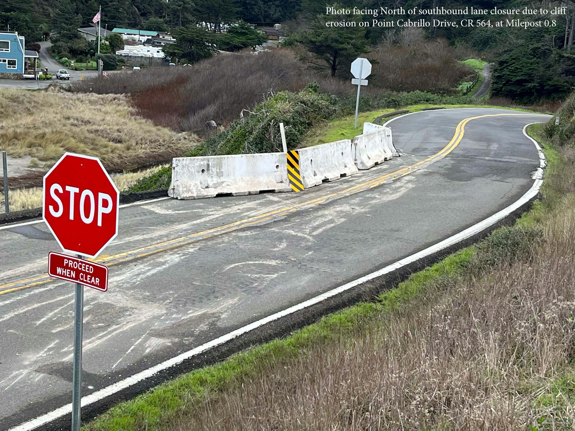 Stop sign, lane closure with barriers on a road surrounded by grass and trees; village buildings and beach sand in distance. Erosion issue noted.