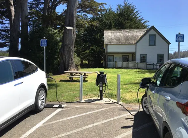 Two electric cars are charging in a lot near a picnic area and a white house, with trees and blue sky in the background.