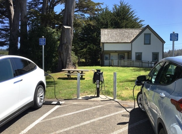 Two electric cars are charging in a lot near a picnic area and a white house, with trees and blue sky in the background.